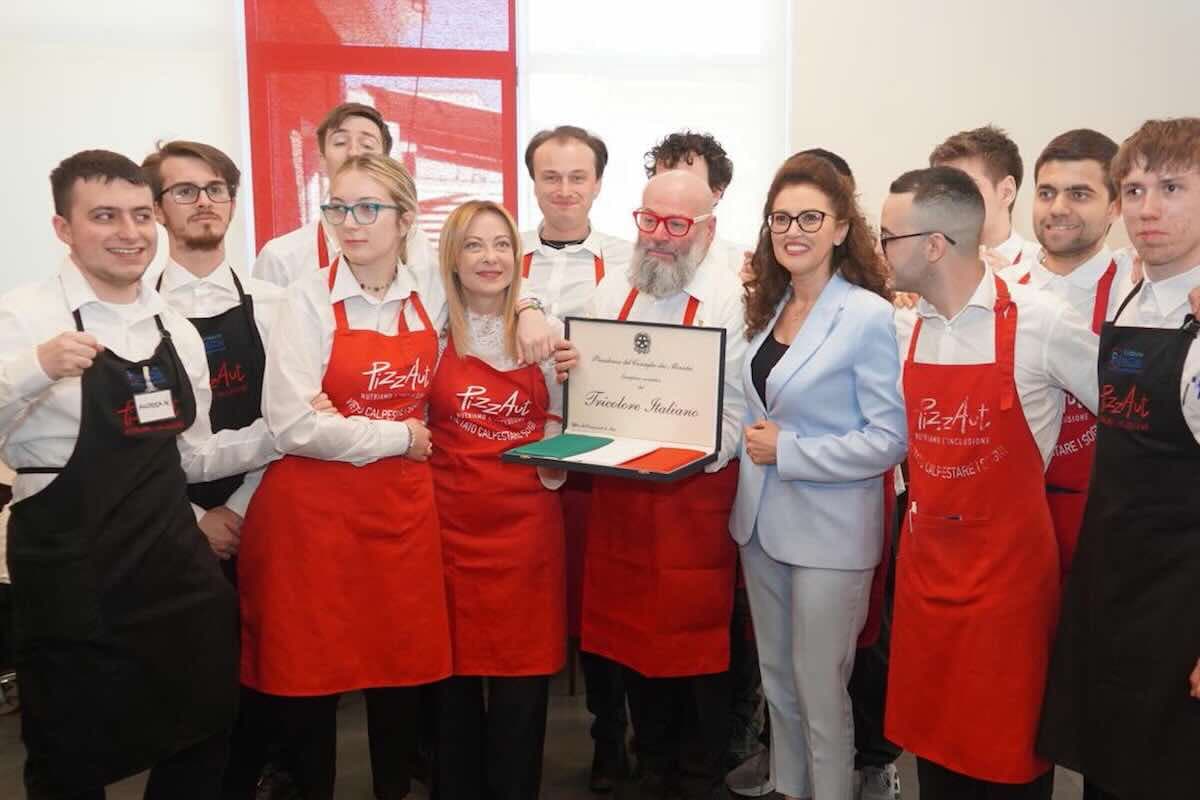 Meloni a pranzo dai ragazzi di PizzAut per il Primo Maggio, la firma delle promozioni e il grembiule rosso in dono: «L’Italia è orgogliosa di voi»